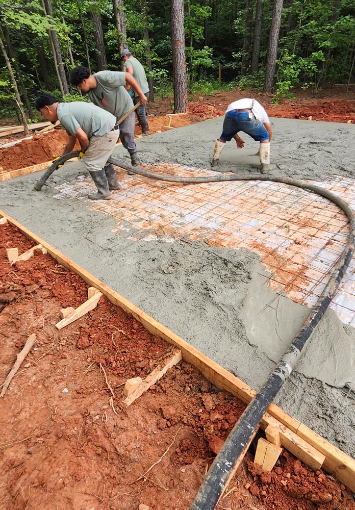 Three workers pouring and spreading concrete over a rebar grid on a construction site surrounded by trees.