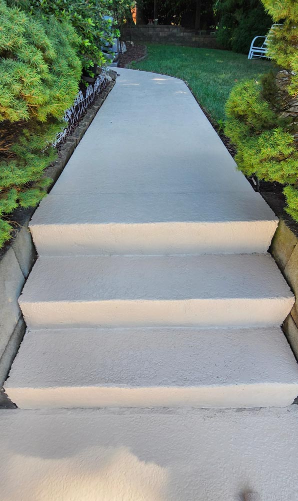 Concrete steps leading up to a smooth path bordered by greenery, with grass and a white chair in the background.