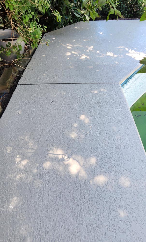 Concrete poolside deck with textured surface, dappled sunlight, and leafy green plants nearby.