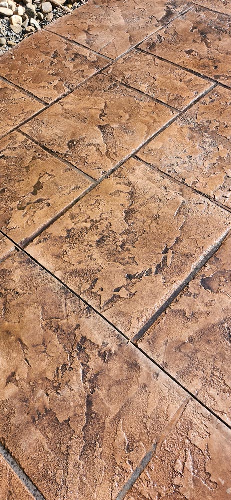Stamped concrete patio with a textured, brown pattern resembling natural stone tiles. Rocks visible at the edge.