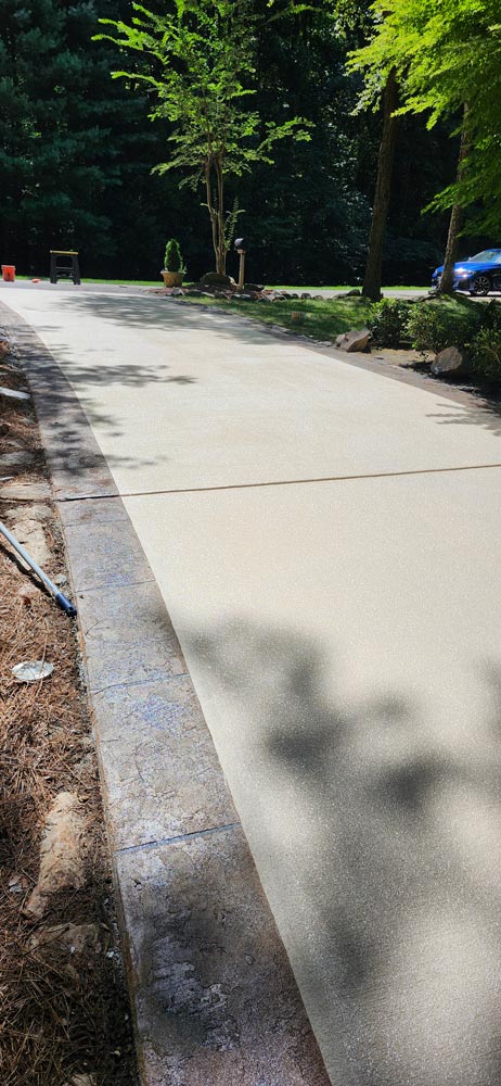Freshly paved concrete driveway bordered by stone, surrounded by trees and landscaped greenery.
