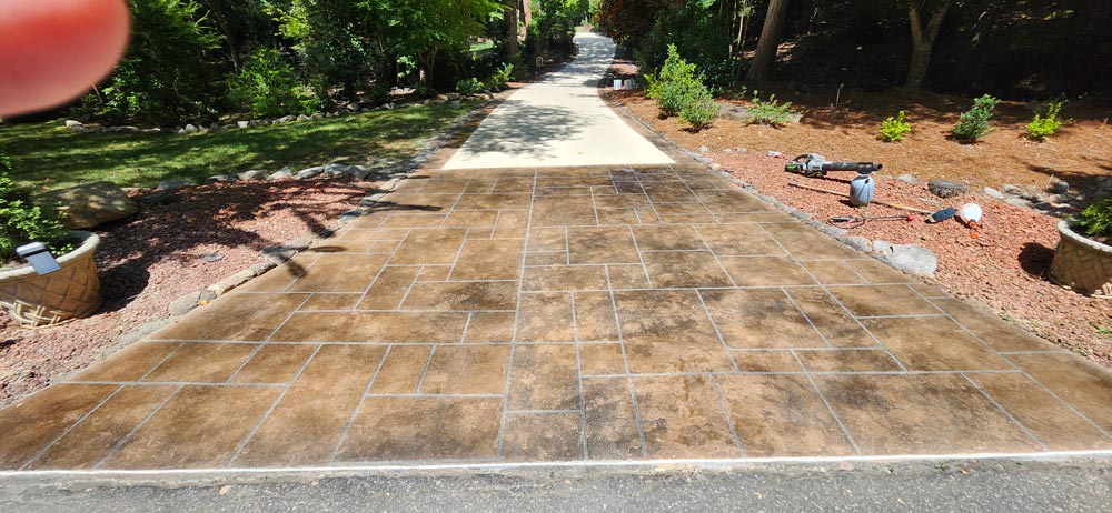 A patterned concrete driveway with a stamped design leads to a shaded, tree-lined path.