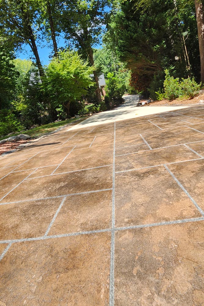 Brown stamped concrete driveway with a grid pattern, surrounded by trees and greenery in sunlight.