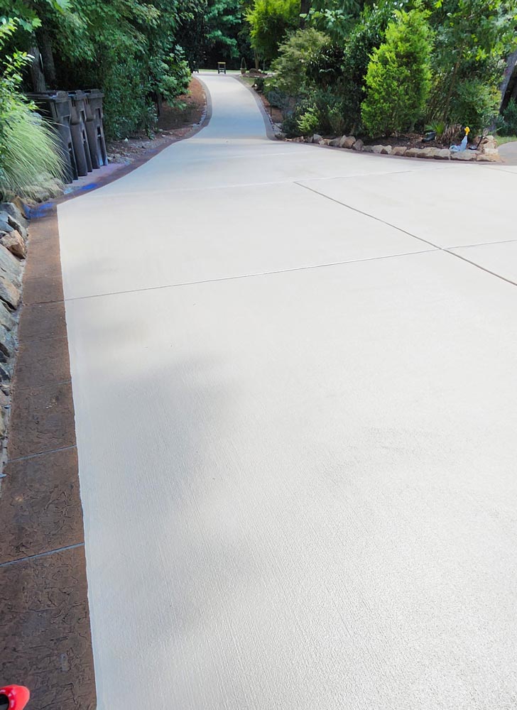 A long, clean concrete driveway curves uphill, lined with green trees and shrubs on both sides.