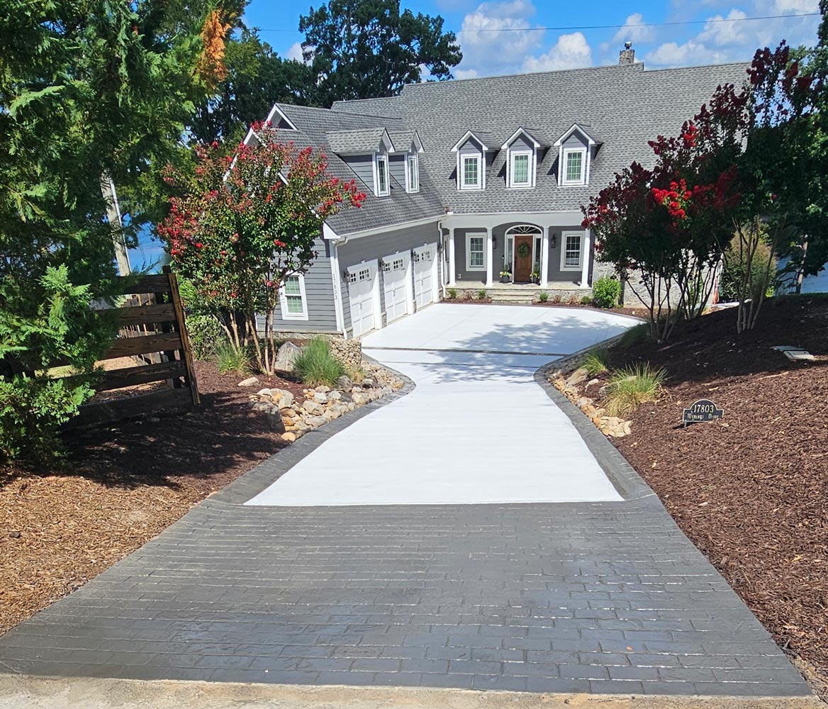 A gray house with dormer windows and a white driveway lined with plants and mulch.