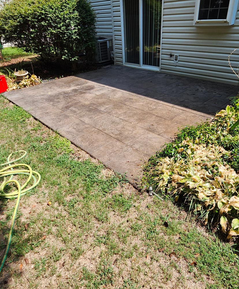 Concrete patio beside a house, bordered by grass, a green hose, and plants with wilted leaves.