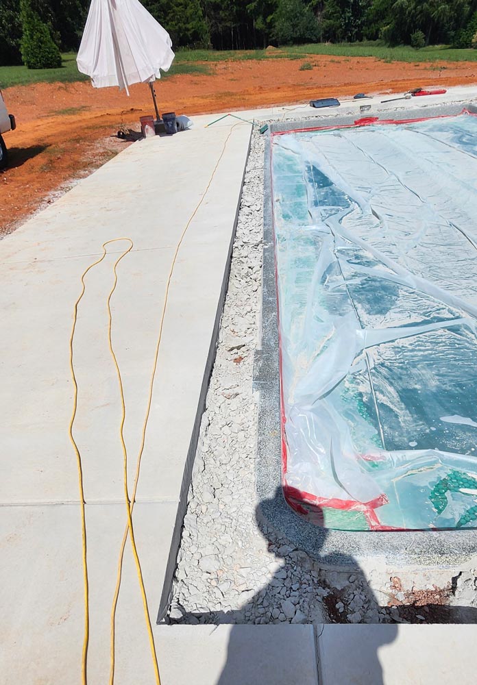 A pool under construction is covered with plastic beside a concrete patio with yellow cords on the ground.