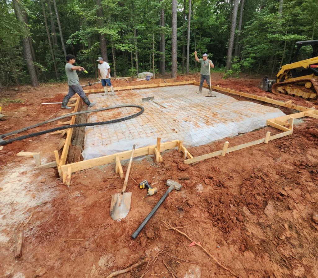 Three workers prepare a concrete foundation at a construction site in a wooded area with tools nearby.