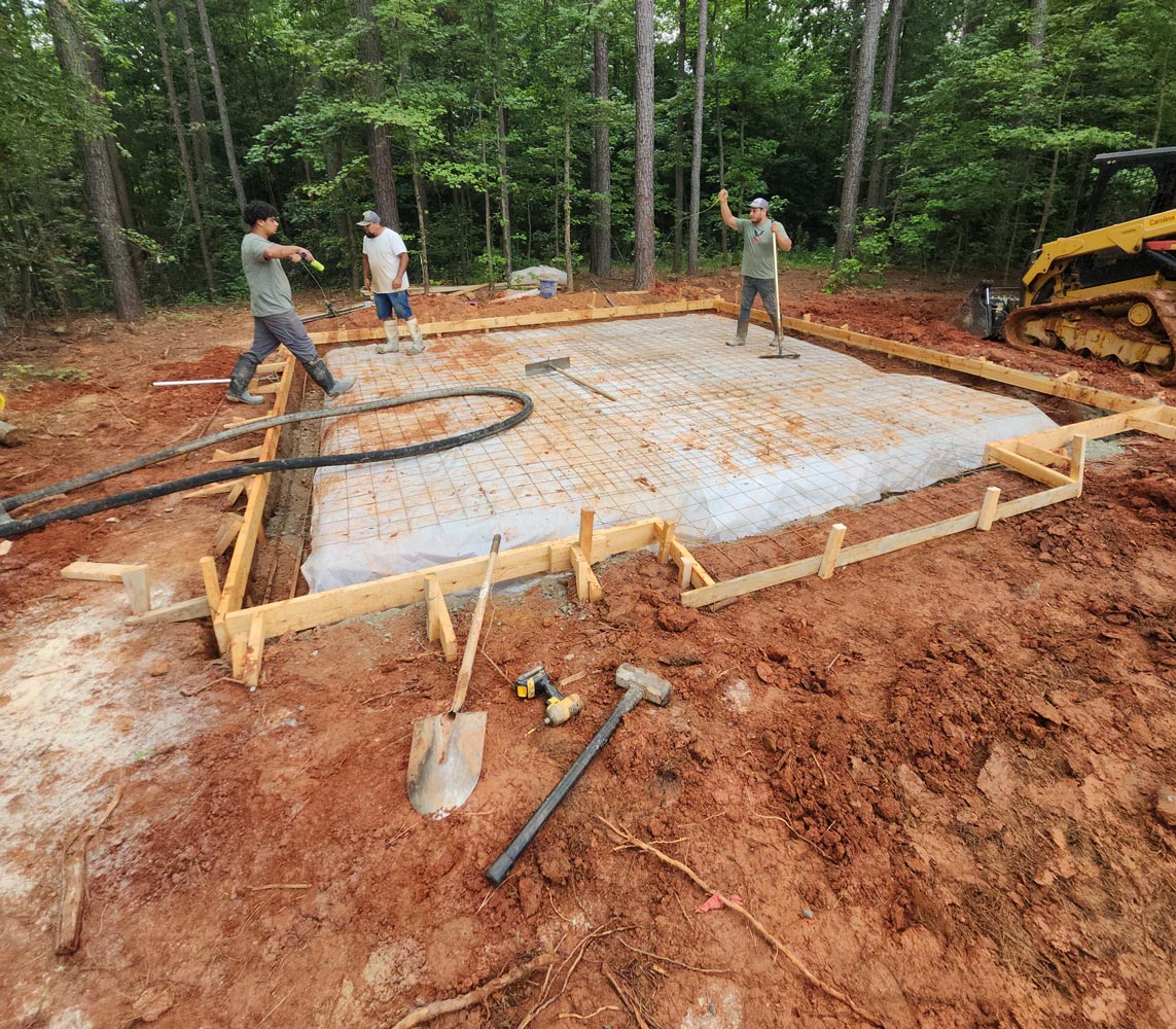 Three workers prepare a concrete slab foundation in a wooded area with construction tools and equipment nearby.