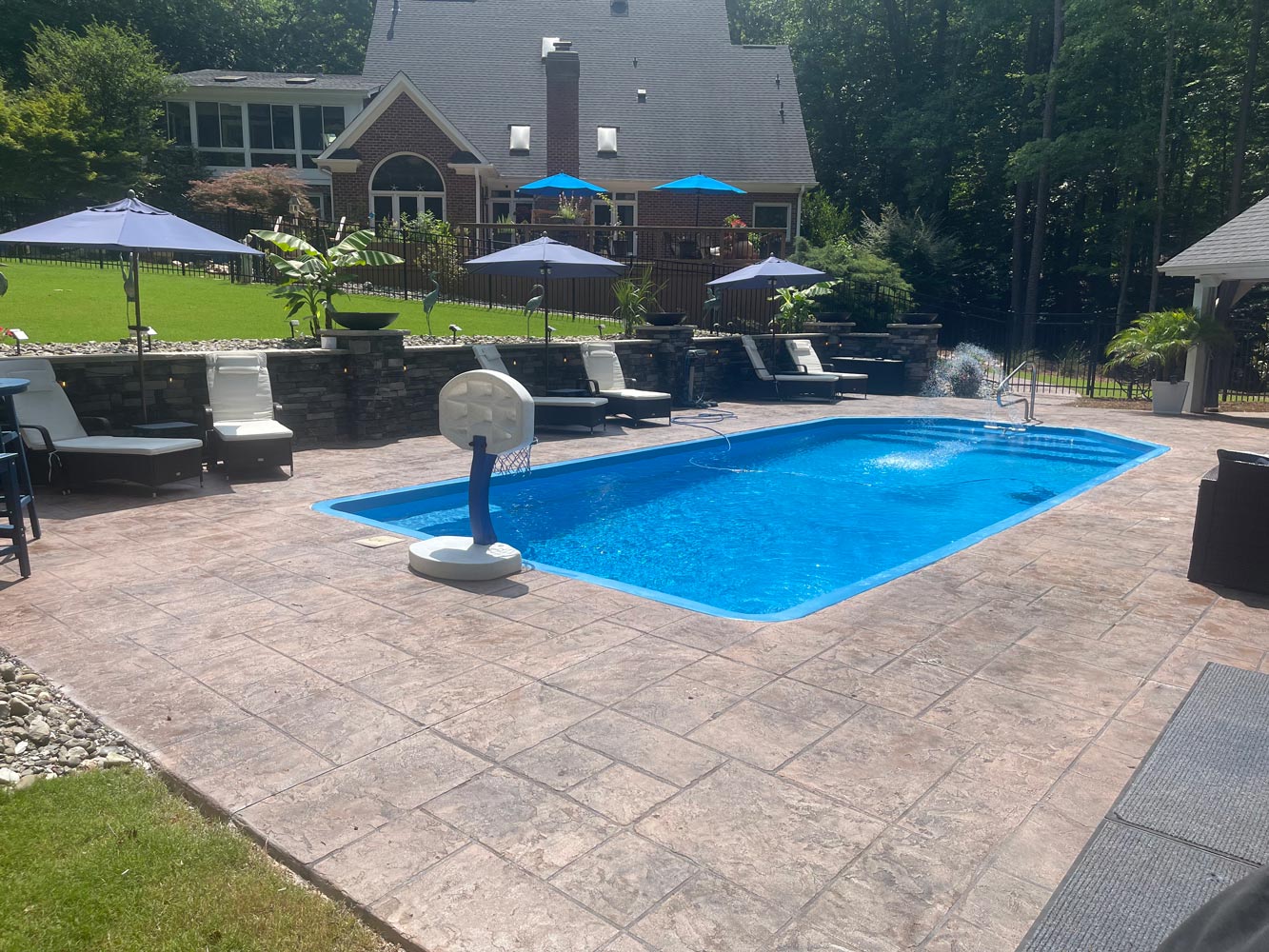 IMG_20250625_143150 A backyard pool with lounge chairs, umbrellas, and a basketball hoop, beside a large house and green lawn.