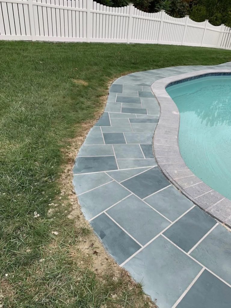 Curved stone pool deck meets lawn, with a white fence in the background and a small patch of bare soil visible.