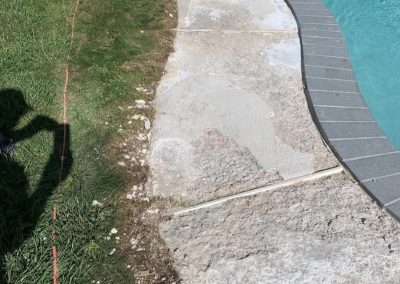 Worn, cracked concrete poolside next to grass with a red cord and white plastic bag on the ground.