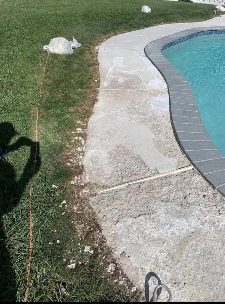 Worn, cracked concrete poolside next to grass with a red cord and white plastic bag on the ground.