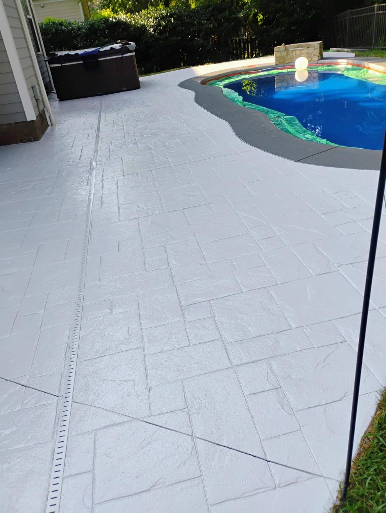White-stamped concrete patio beside a pool with blue water and greenery in the background.