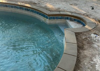 Partially filled pool with clean tiles and water, bordered by dirt and concrete, near a lake and trees.