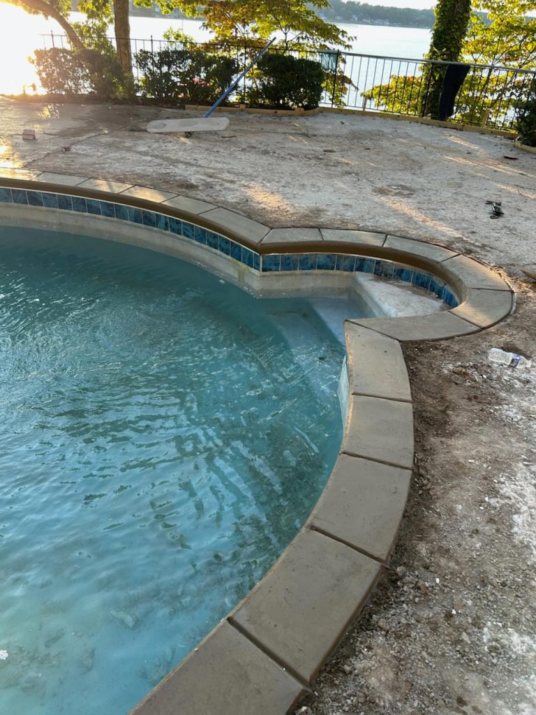 Partially filled pool with clean tiles and water, bordered by dirt and concrete, near a lake and trees.
