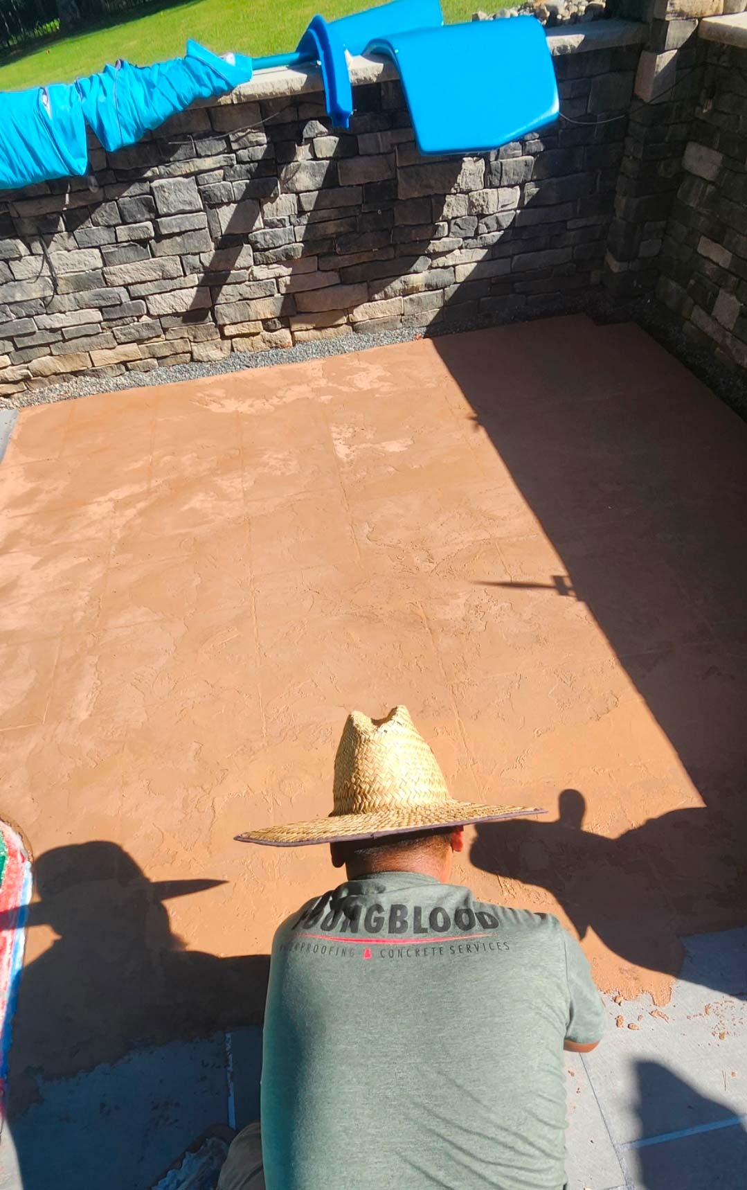 Man in a straw hat smoothing wet concrete on a patio near a stone wall and blue slide.