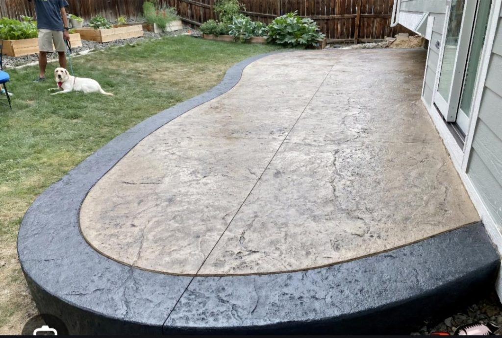Stamped concrete patio with a black border, next to grass; a dog and person stand nearby in the yard.