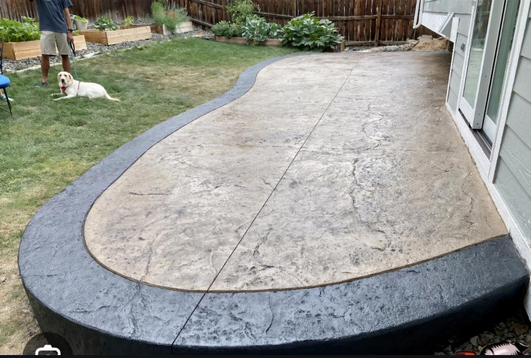 Curved concrete patio with a dark border next to grass; a dog and a person stand in the yard.