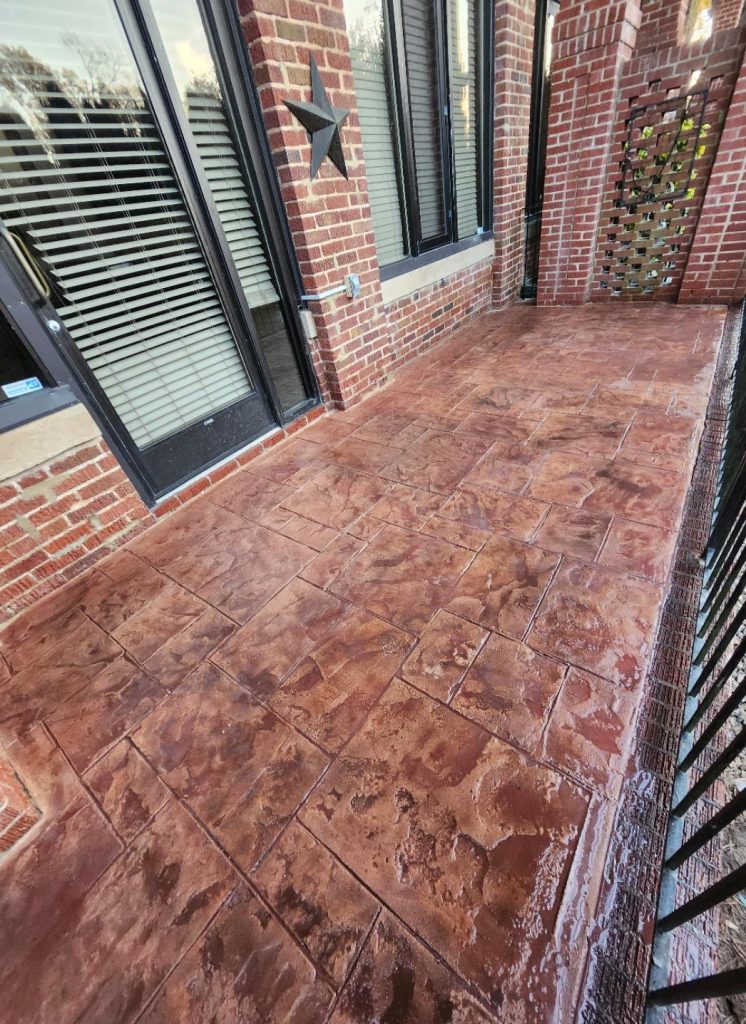 Stamped concrete patio with a wet, reddish surface beside a brick wall and black metal railing.