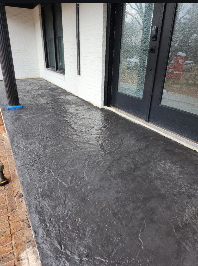 A textured, dark grey concrete porch in front of a house with white walls and black framed windows and doors.