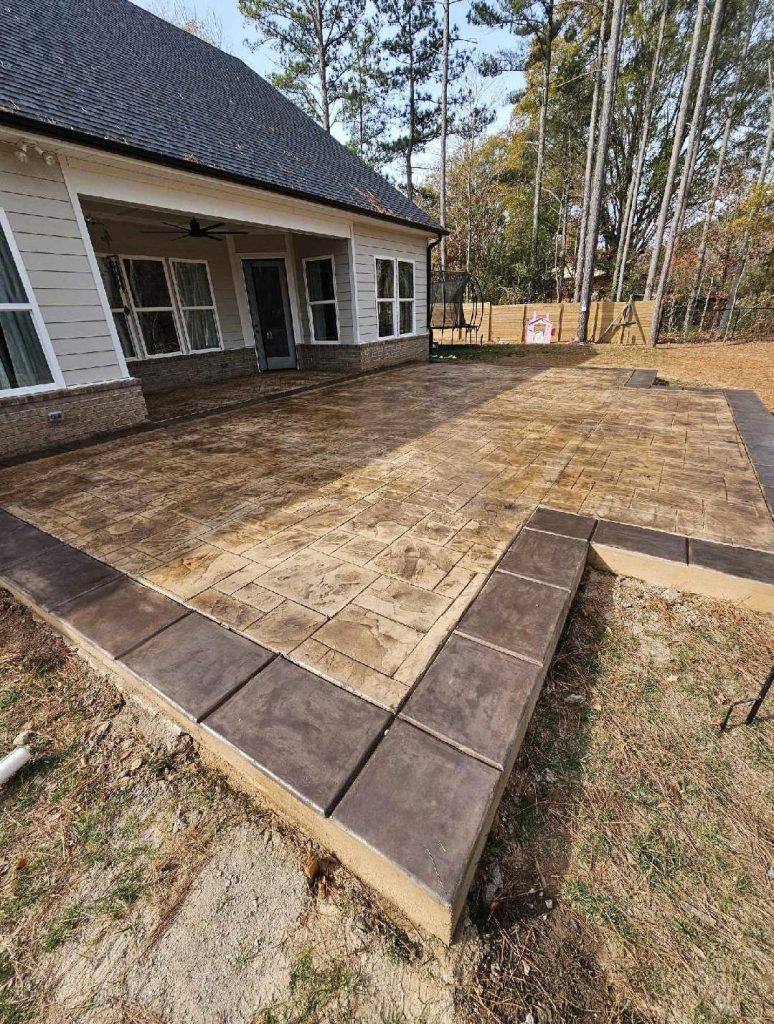 Stamped concrete patio with brown borders beside a house, surrounded by grass and trees in the background.