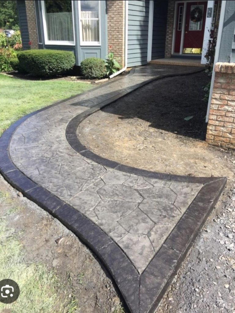 Curved, stamped concrete walkway with dark borders leads to the front door of a house with landscaped yard.