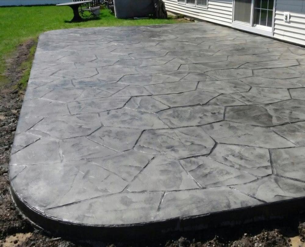 Stamped concrete patio with a stone pattern next to a house, surrounded by grass and soil.