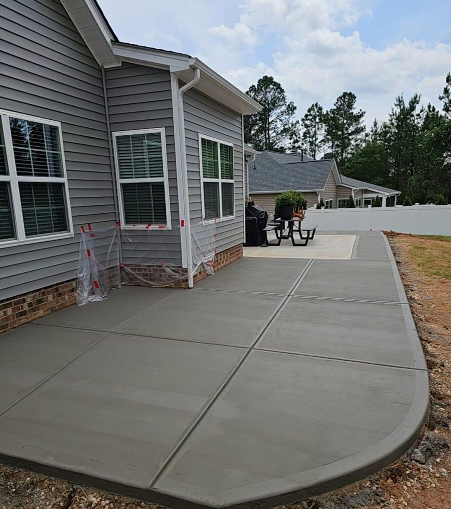 Freshly poured concrete patio beside a gray house with outdoor furniture and grill in the background.