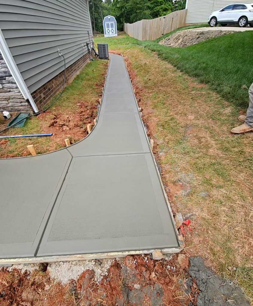 Freshly poured concrete sidewalk next to a house, with grass and tools visible on the side.
