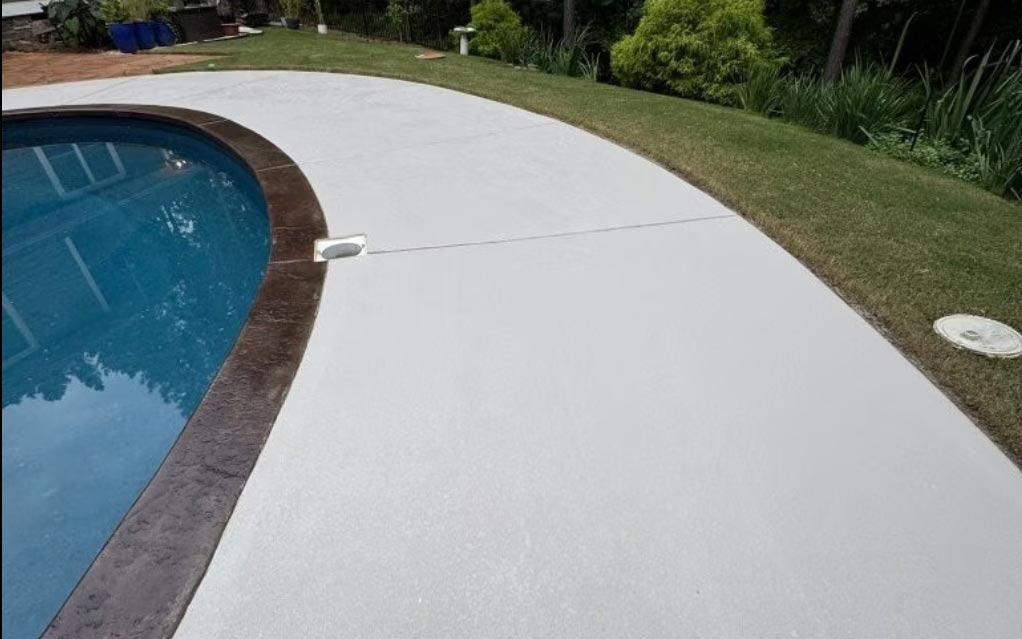 A clean, smooth concrete pool deck curves beside a blue swimming pool and green grass.