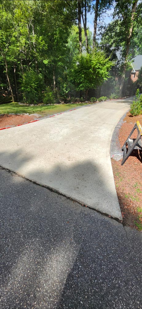 A newly paved concrete driveway surrounded by trees and landscaping on a sunny day.
