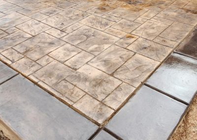 Stamped concrete patio with a decorative border next to a house and some exposed soil in the foreground.