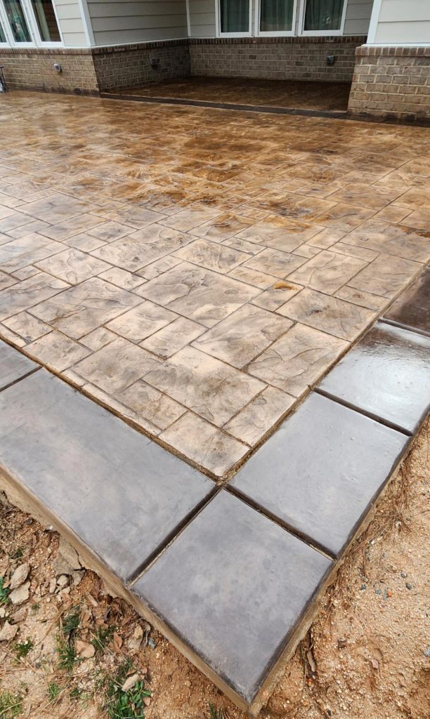 Stamped concrete patio with a decorative border next to a house and some exposed soil in the foreground.