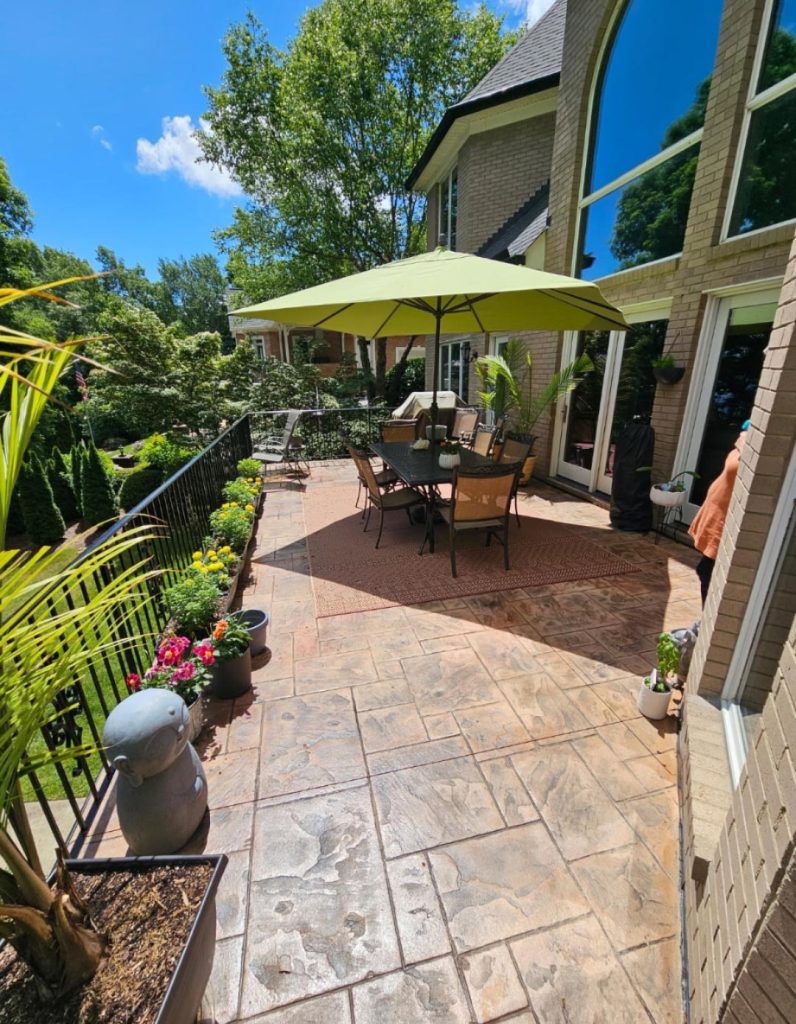 Sunny patio with table, chairs, and green umbrella, surrounded by potted plants and a landscaped garden.