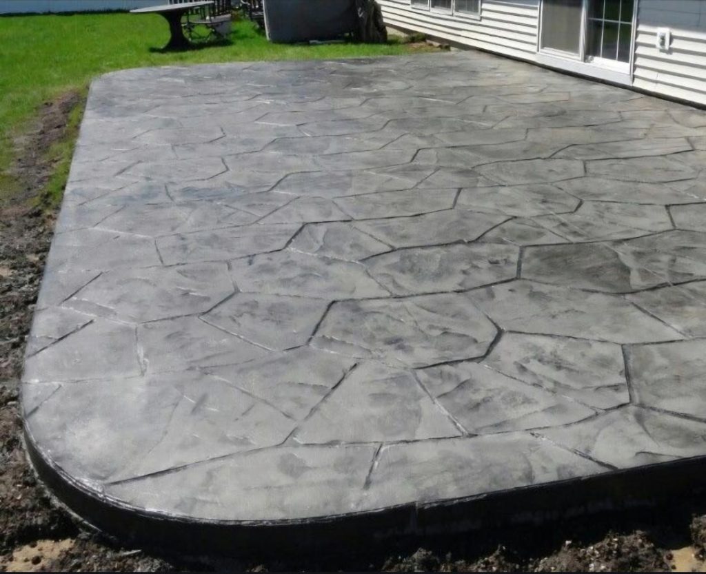 Stamped concrete patio with a stone pattern next to a house, surrounded by grass and dirt.