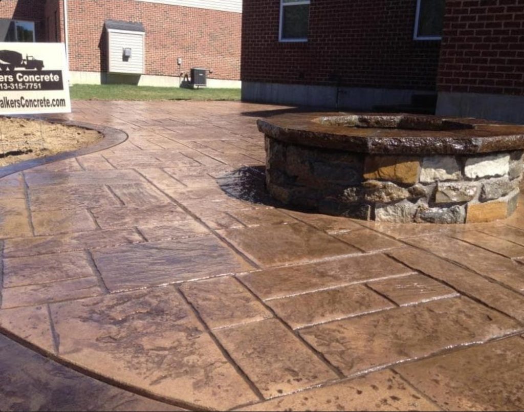 Stamped concrete patio with a round stone fire pit in a backyard next to a brick building.