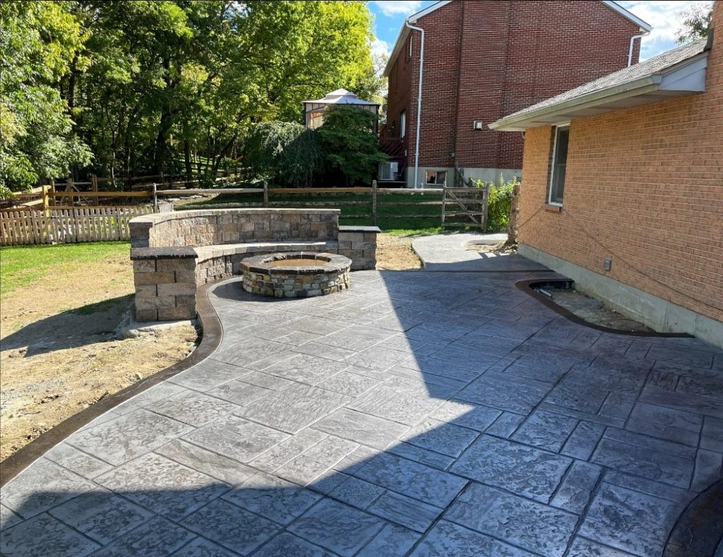 Stamped concrete patio with a round stone fire pit and curved stone seating wall beside a brick house.