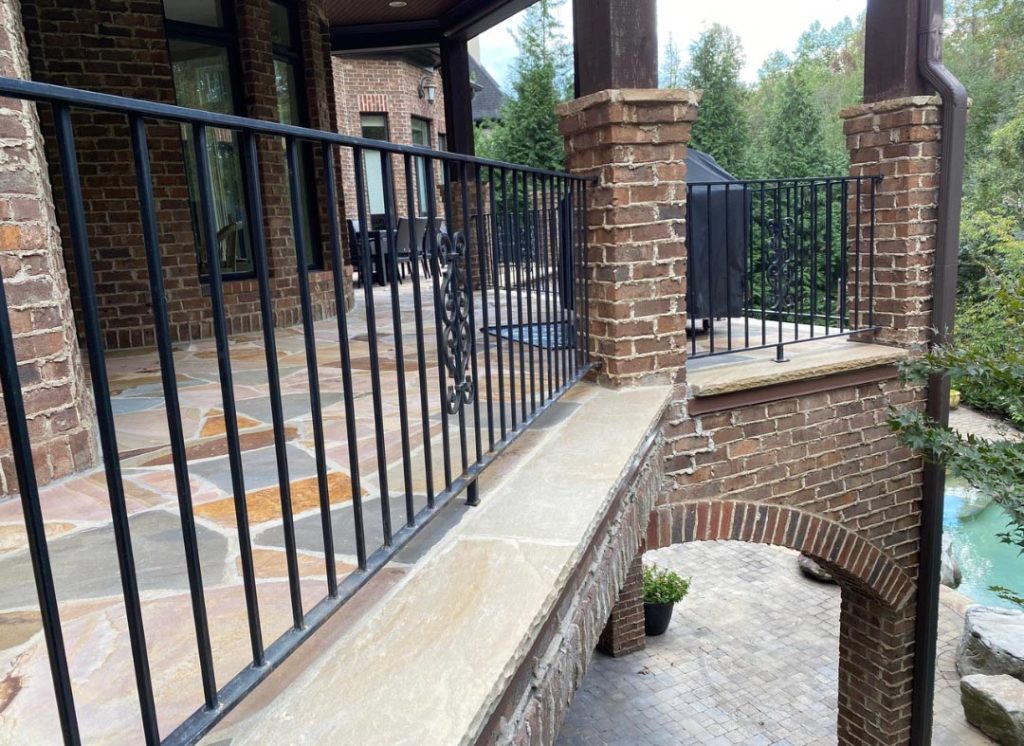 Brick patio with black metal railings, stone flooring, and an archway overlooking a pool and trees.