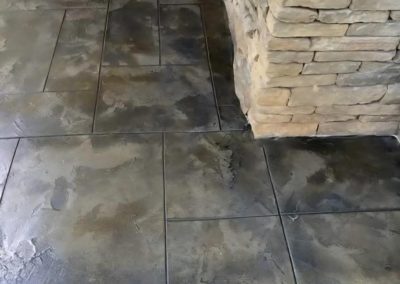 Textured concrete floor with large rectangular slabs next to a corner of a stacked stone wall.
