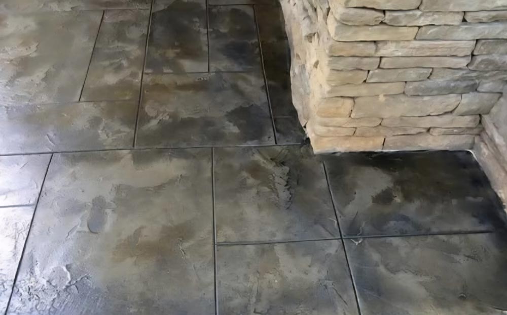 Textured concrete floor with large rectangular slabs next to a corner of a stacked stone wall.