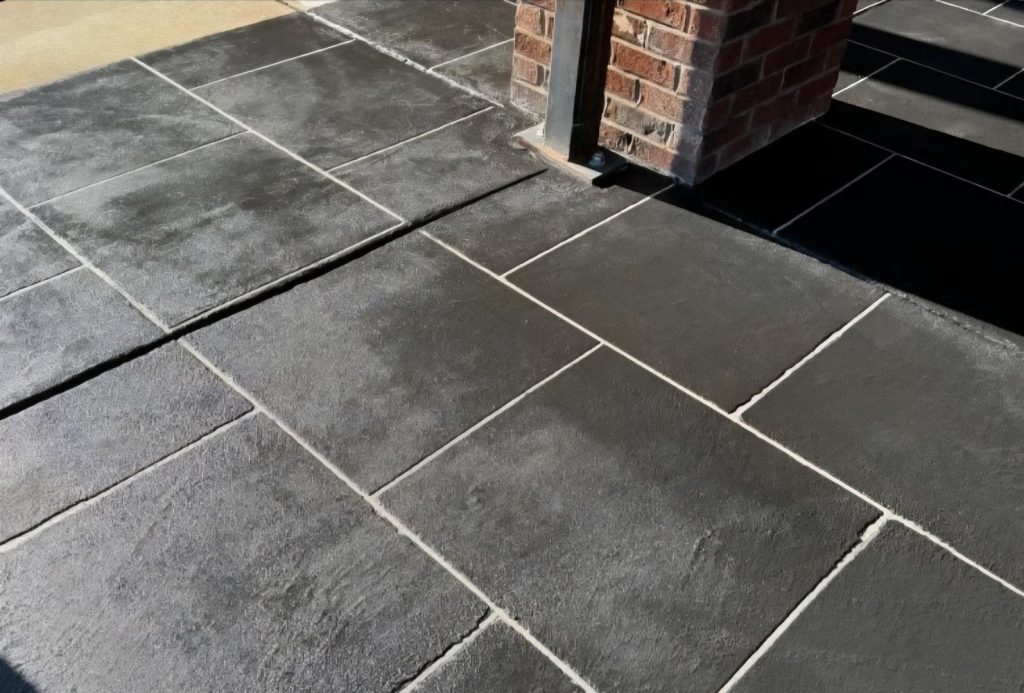 Gray concrete tiles with light grout lines form a patio around a brick and metal post in sunlight.