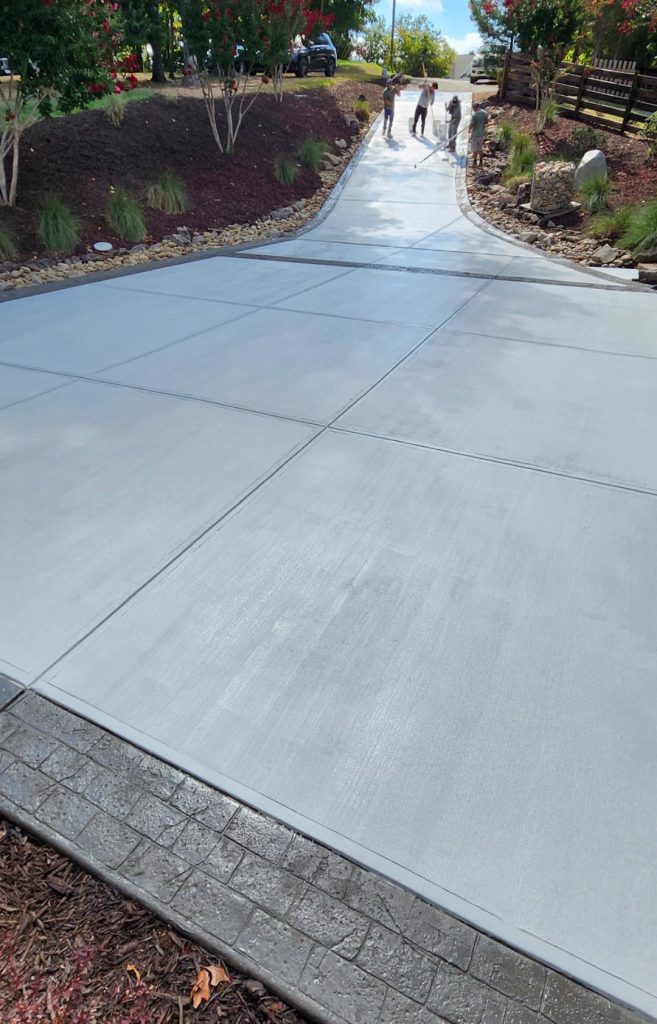 Freshly poured concrete driveway with three people working at the top near landscaped garden beds.