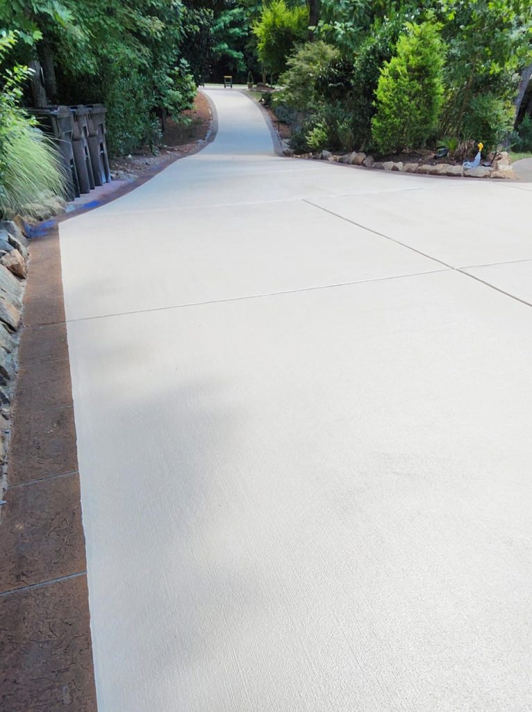 A long, clean concrete driveway lined with greenery and trees on both sides.