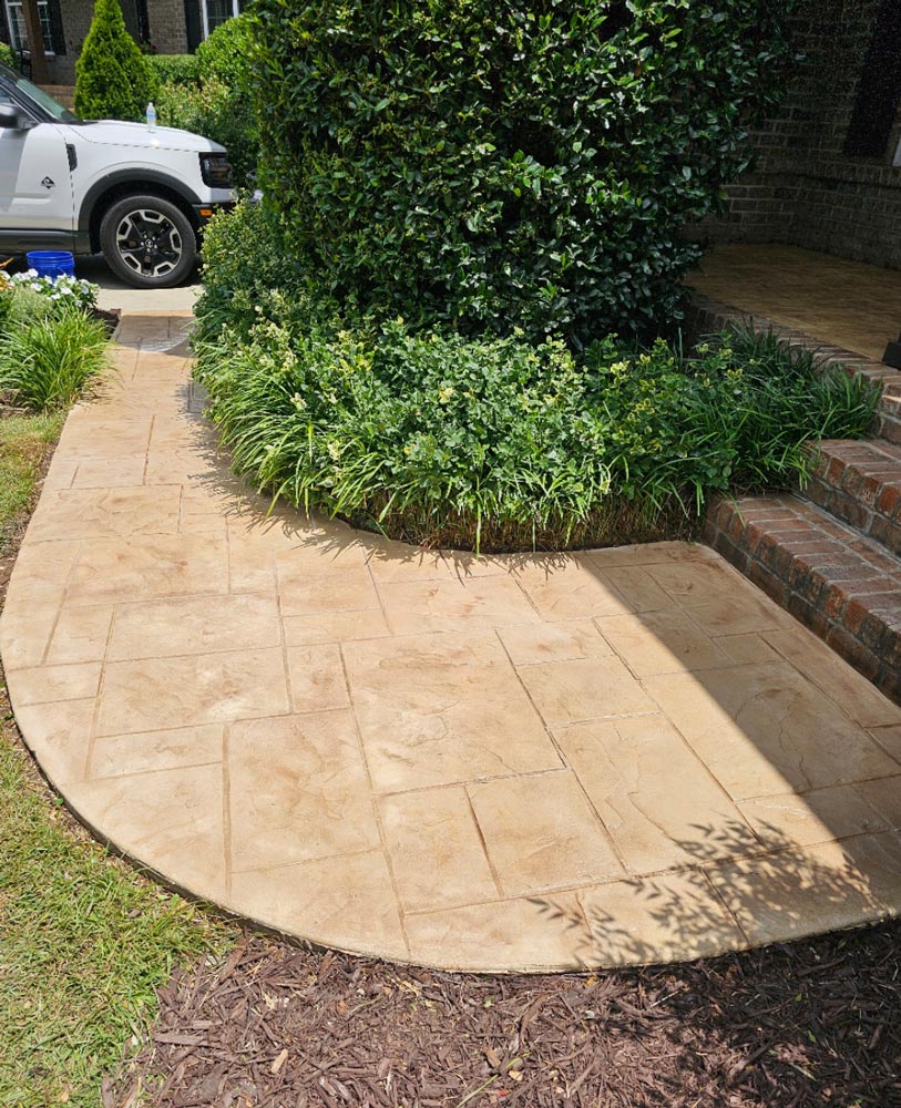 Curved stamped concrete walkway by a bushy garden bed leading to brick steps and a white vehicle.