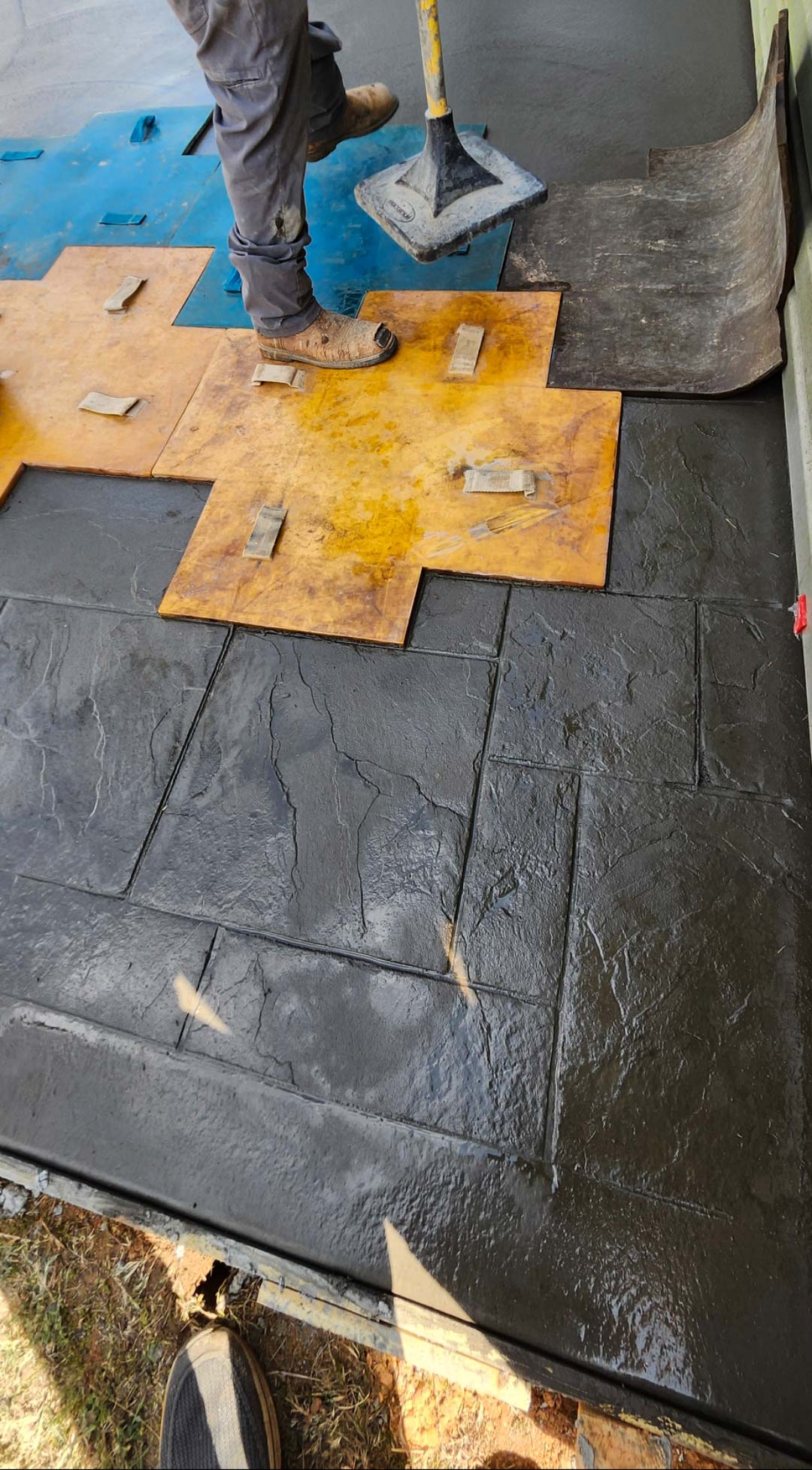 A worker stamps a pattern onto wet concrete using large textured mats at a construction site.