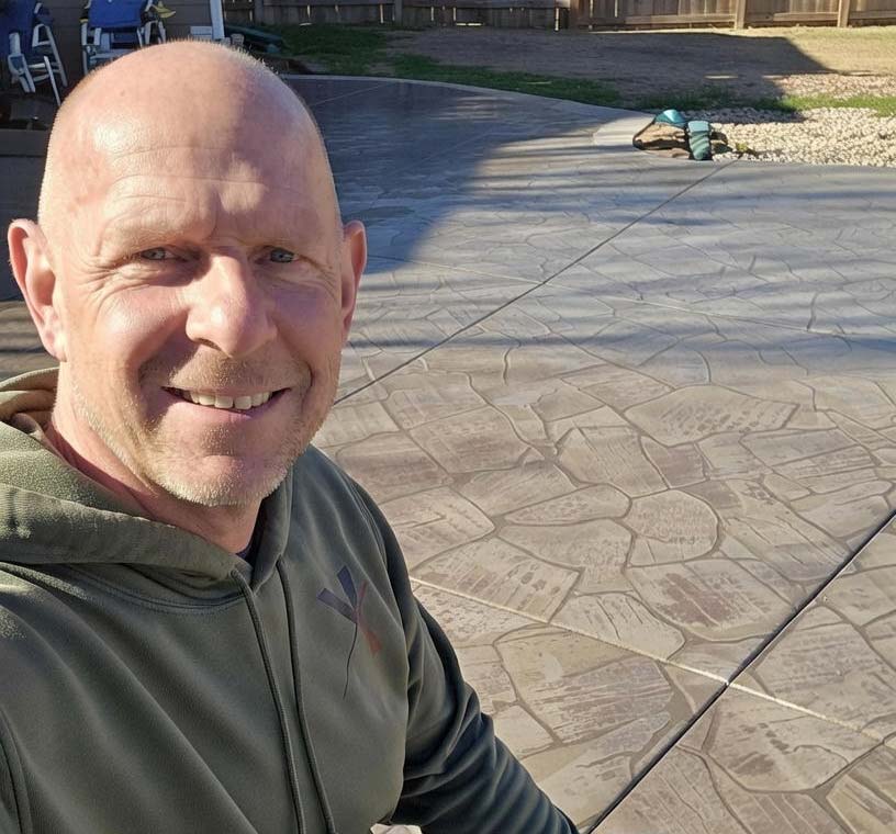 Smiling man in a green hoodie taking a selfie outside on a patterned concrete patio in sunlight.